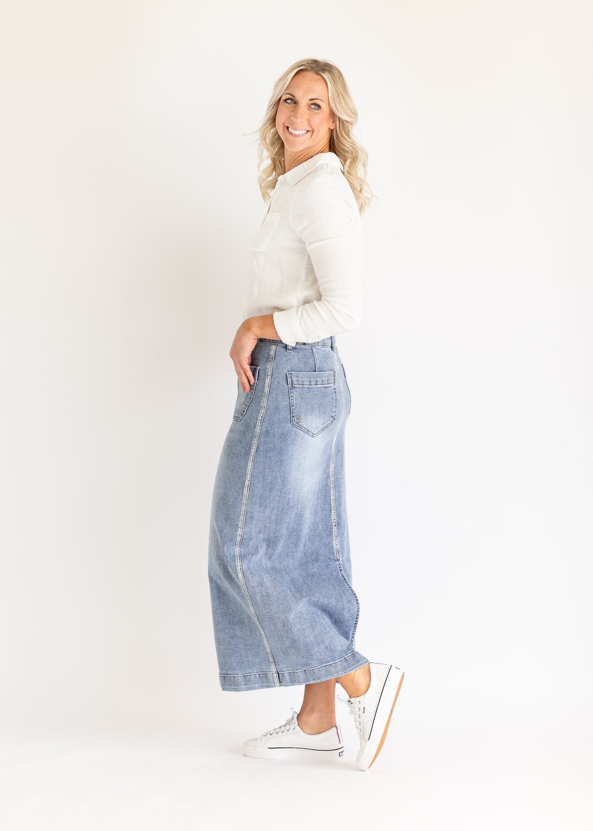 A smiling person stands sideways in a white shirt and long denim skirt, wearing white sneakers. The background is plain white, creating a bright, casual feel.