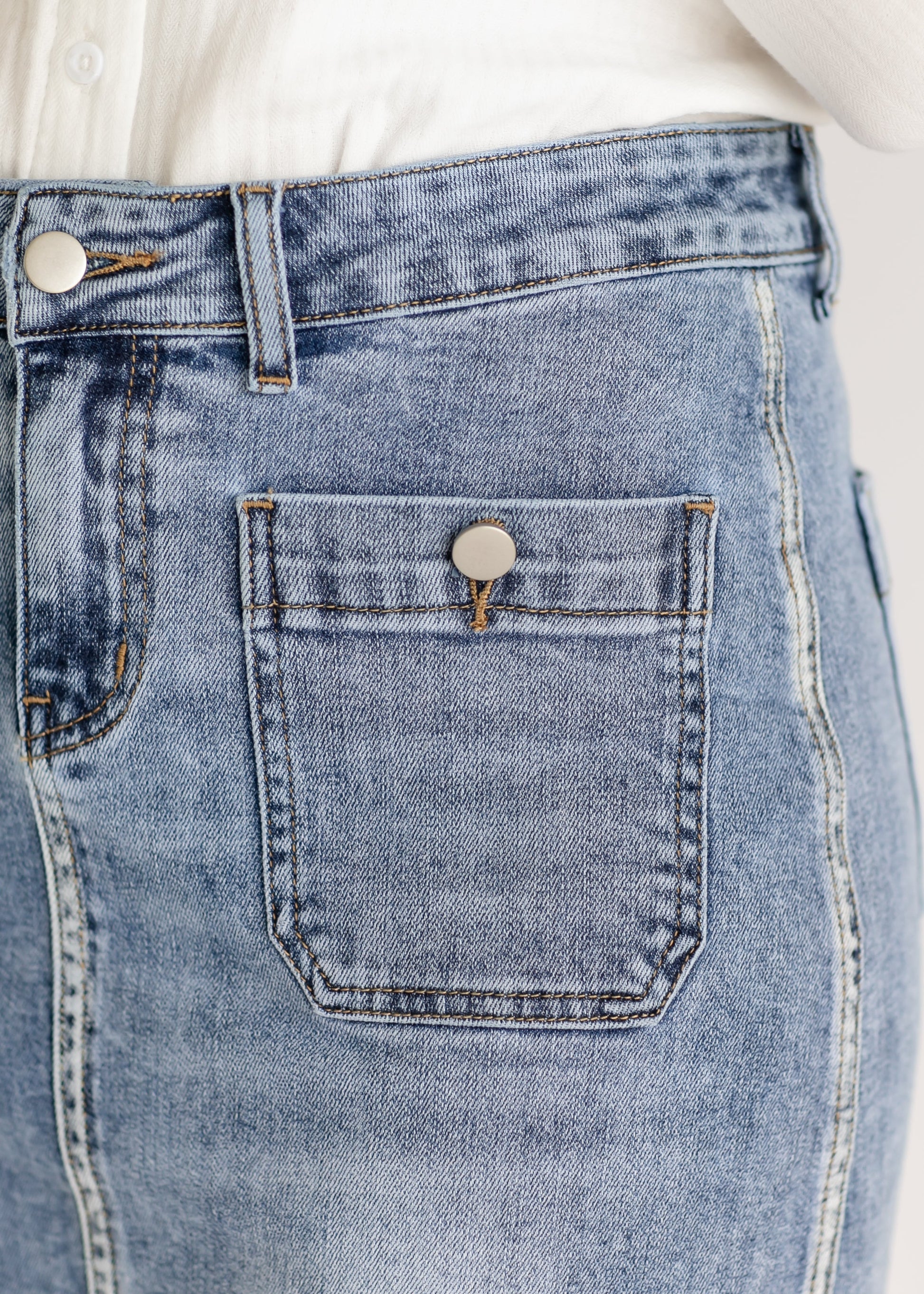 Close-up of light blue denim jeans showing a metal button on the front pocket. The jeans are paired with a white shirt. Casual and classic style.