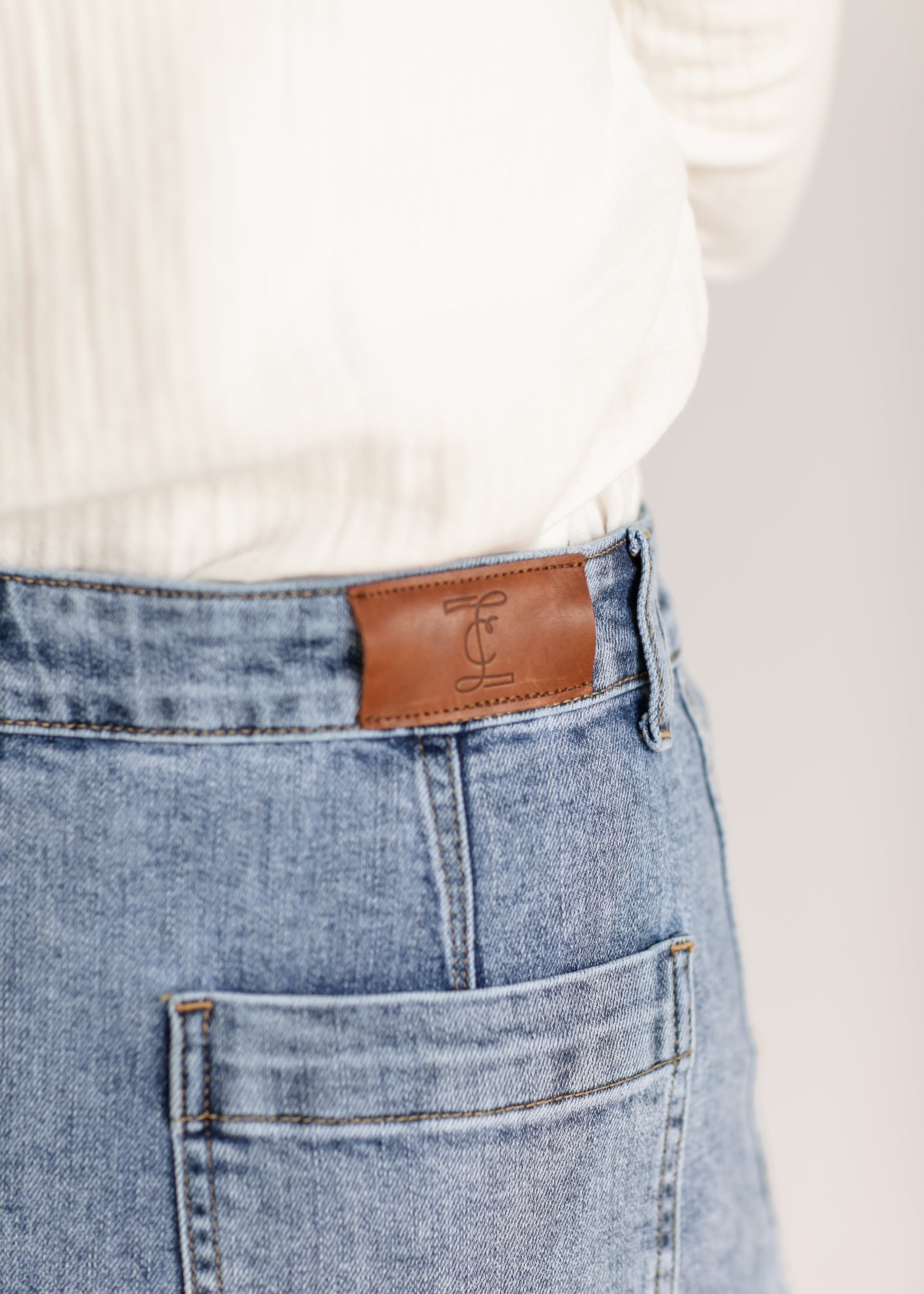 Close-up of the back of a person's light blue denim jeans with a brown leather patch. The person wears a white shirt tucked in, conveying a casual style.