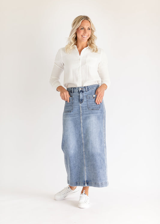 Woman in a white blouse and denim skirt smiles confidently against a plain background. She stands with hands in pockets, wearing white sneakers.