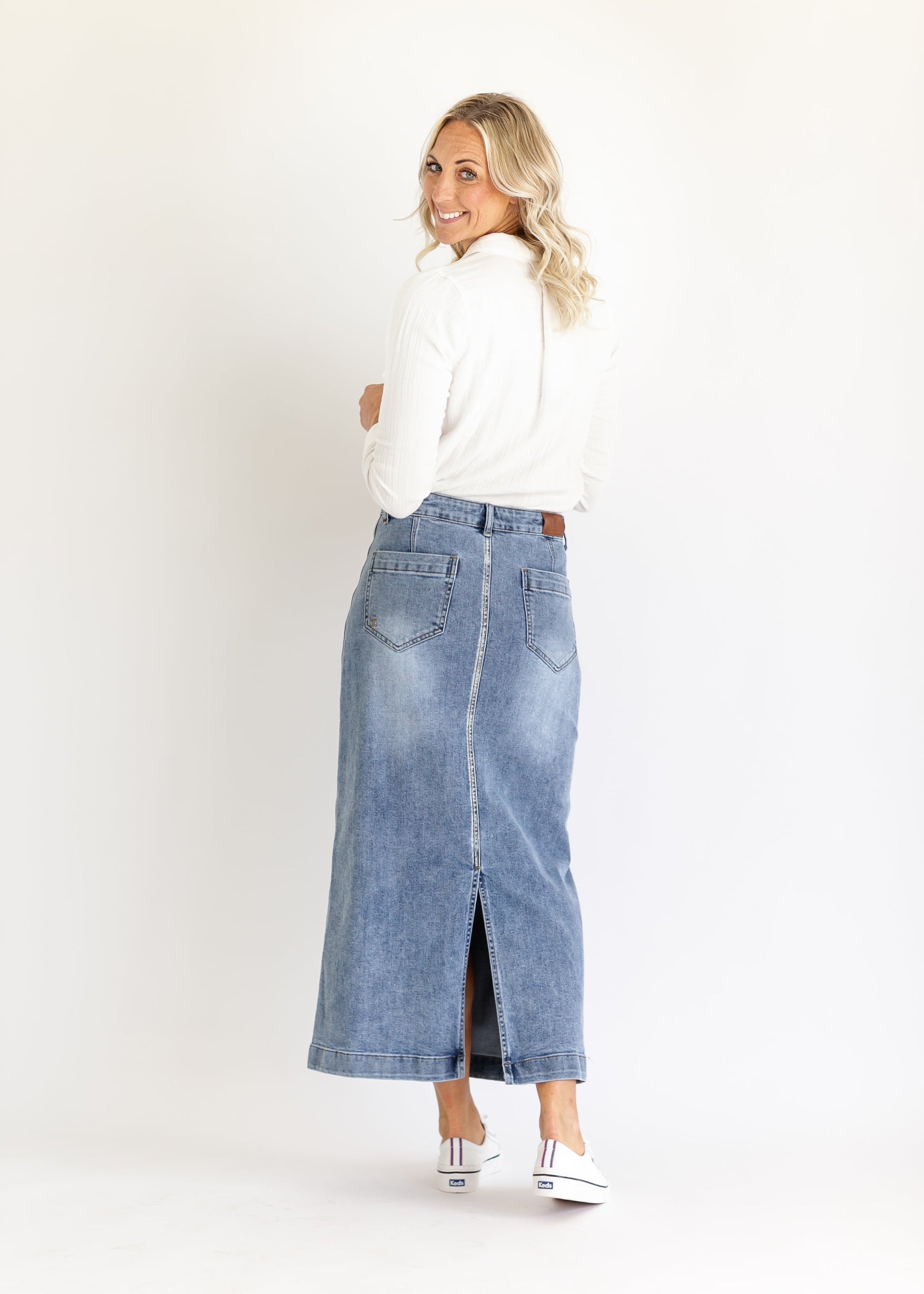 Smiling woman with long blonde hair, wearing a white sweater and long denim skirt, standing against a white background, conveys a casual, cheerful vibe.