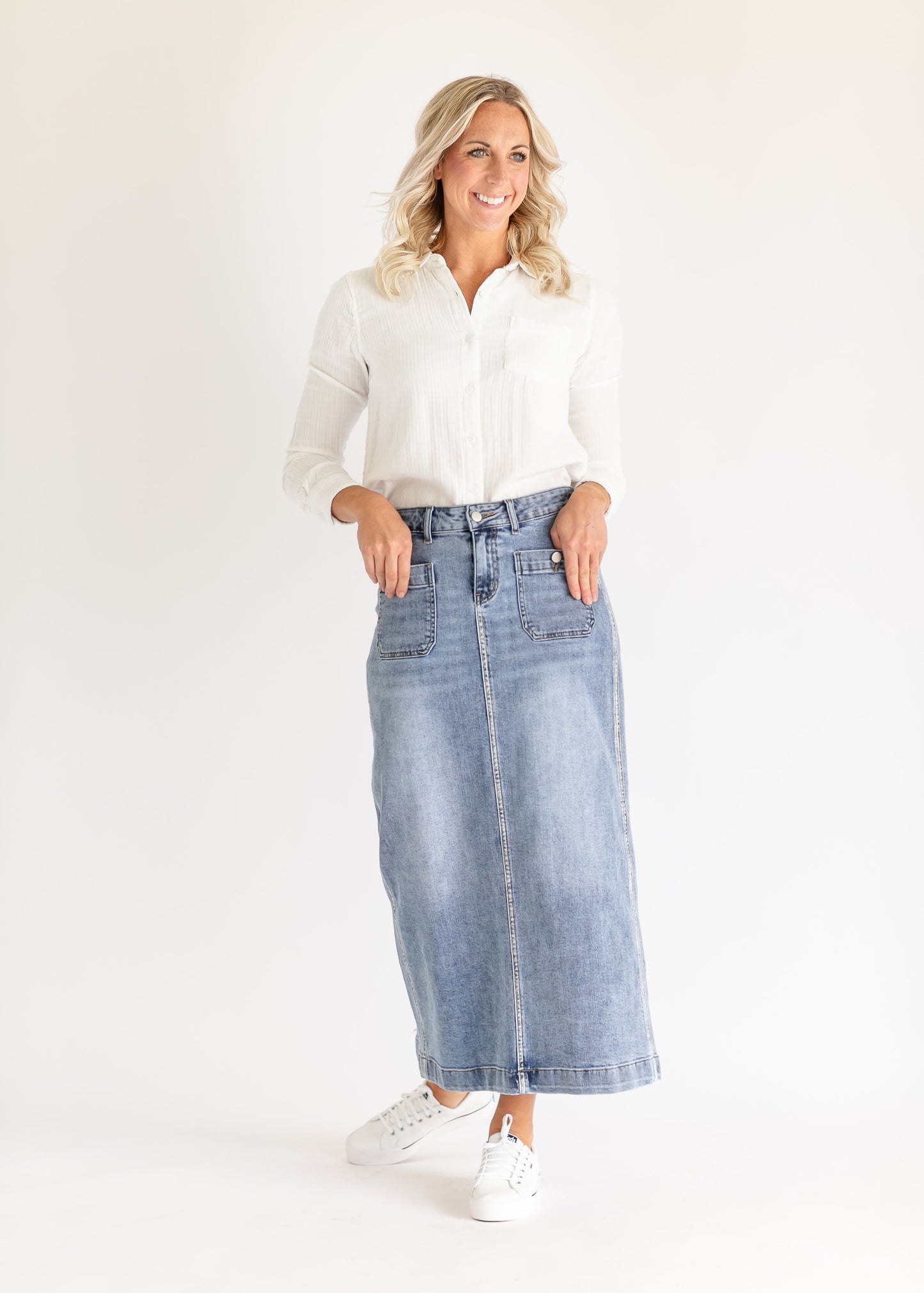 Smiling woman in white blouse and denim skirt stands against a white background, hands in pockets, exuding a casual and positive vibe.