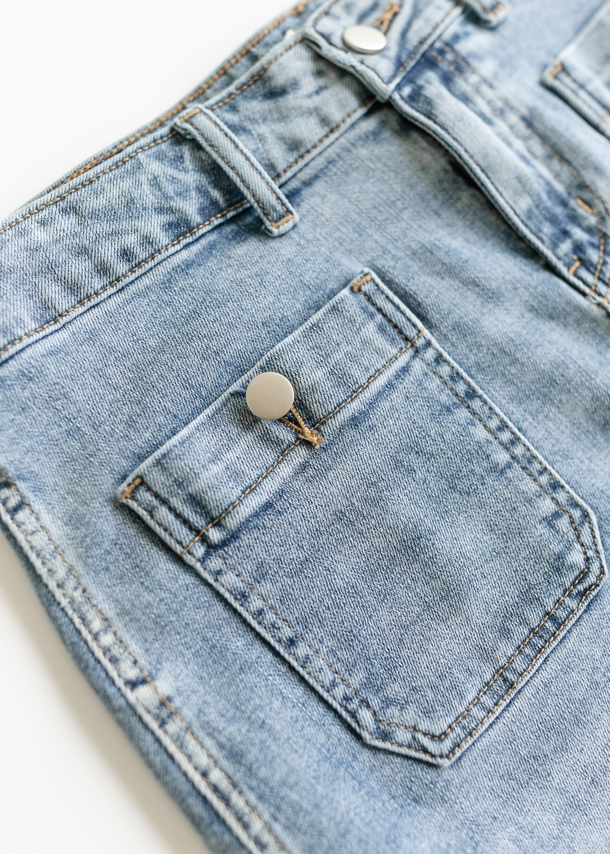 Close-up of light blue denim shorts showcasing a front pocket with a silver button. The stitching is visible, giving a classic and casual look.