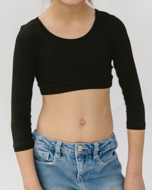 A child wearing a black long-sleeve crop top and blue jeans stands against a white background. The casual outfit suggests a relaxed, everyday setting.