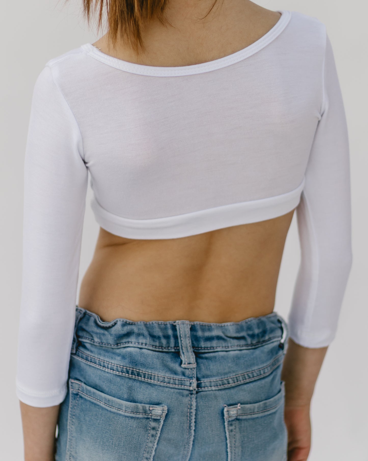 Back view of a person wearing a white, open-back top with three-quarter sleeves and high-waisted blue jeans. The mood is casual and relaxed.