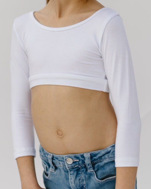 Child wearing a white long-sleeve crop top and blue jeans, standing against a plain background. The pose is relaxed and casual.
