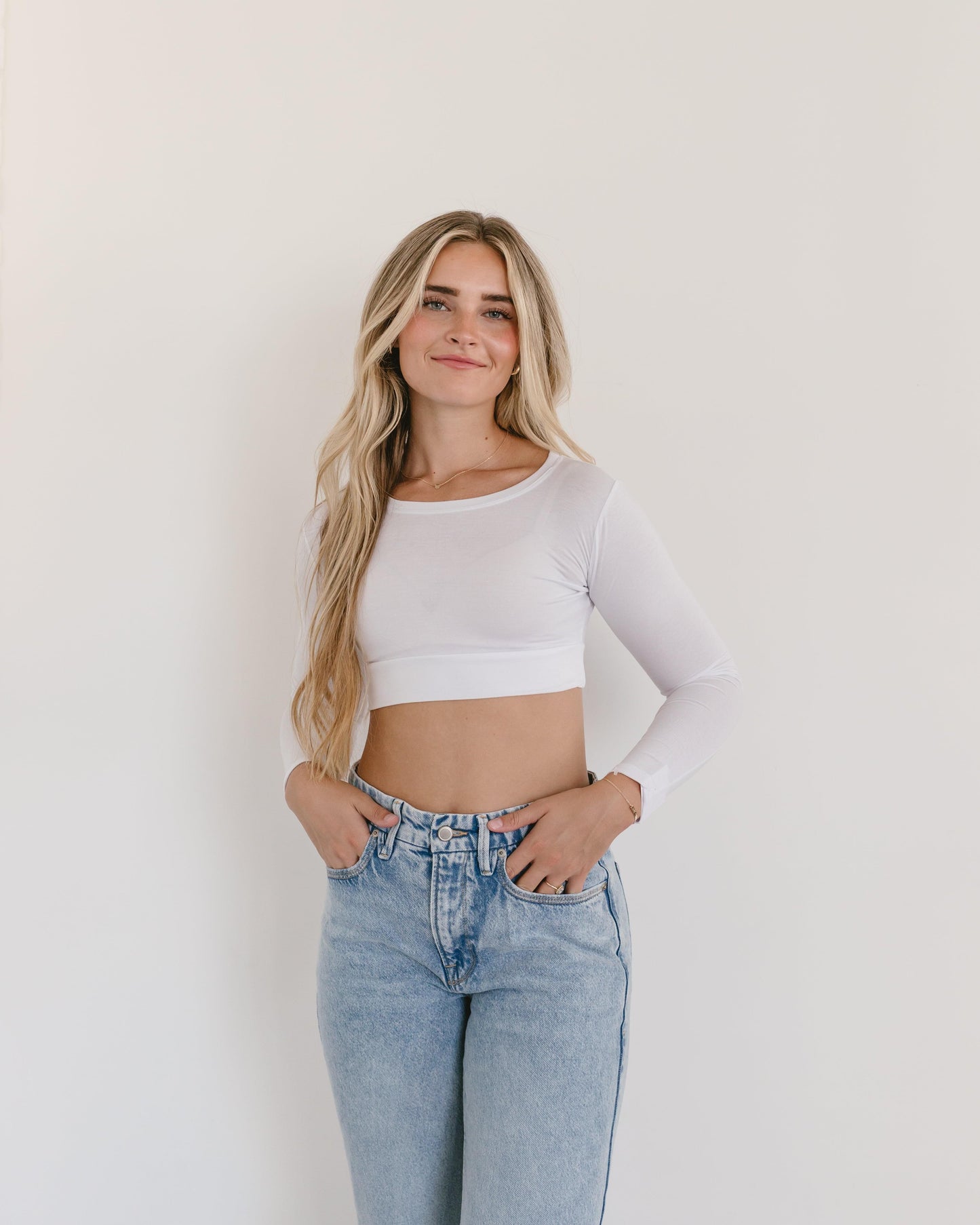 A woman wearing a white crop top and jeans, with a crew neck long sleeve.