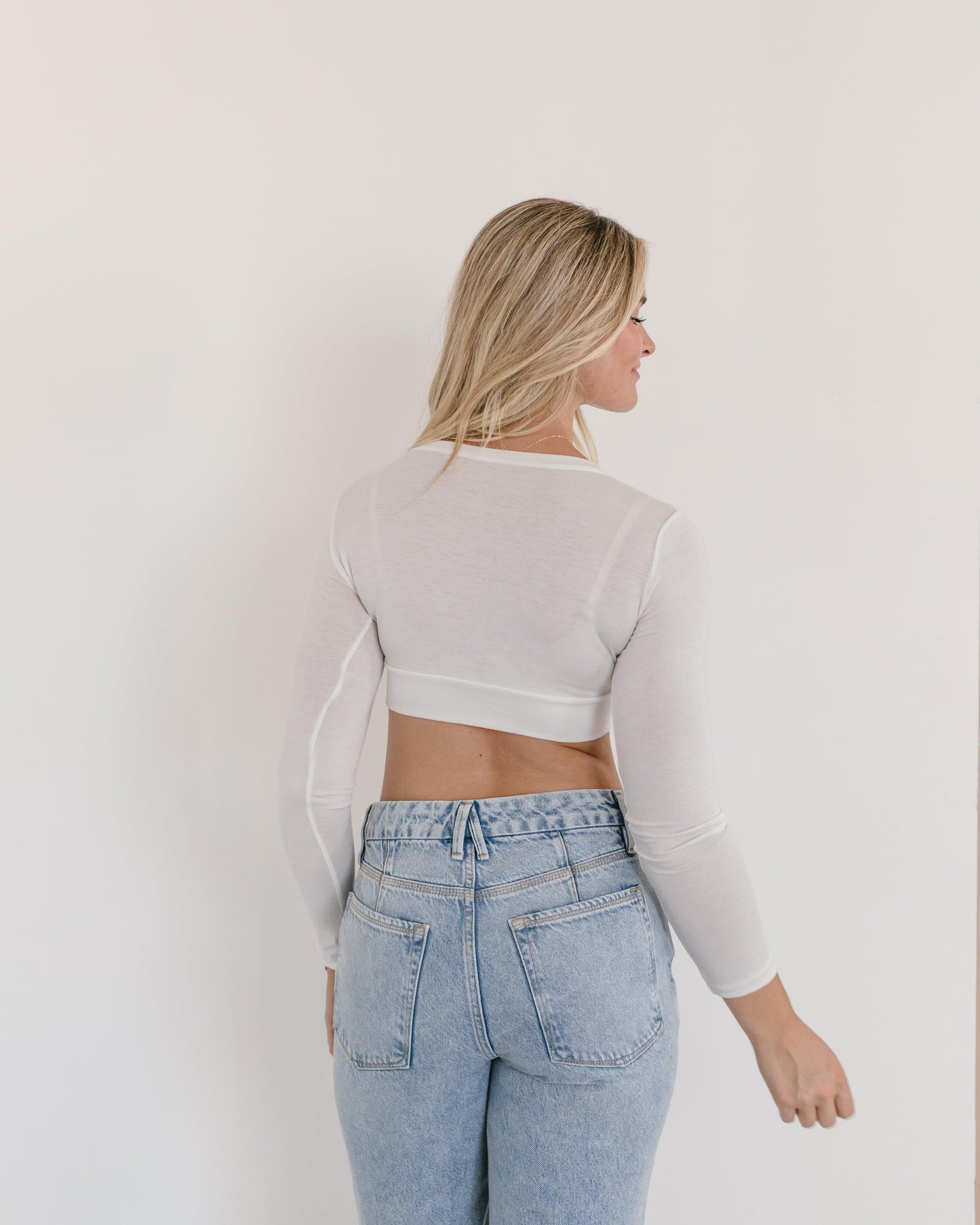 Crew neck long sleeve bralette.  A higher neckline and long sleeves! WhaA woman wearing a white crop top and jeans, with a crew neck long sleeve.t more could a girl want!