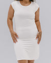 A woman wearing a fitted white dress stands confidently. Her hand rests on her hip, displaying a tattoo. The image conveys simplicity and elegance.