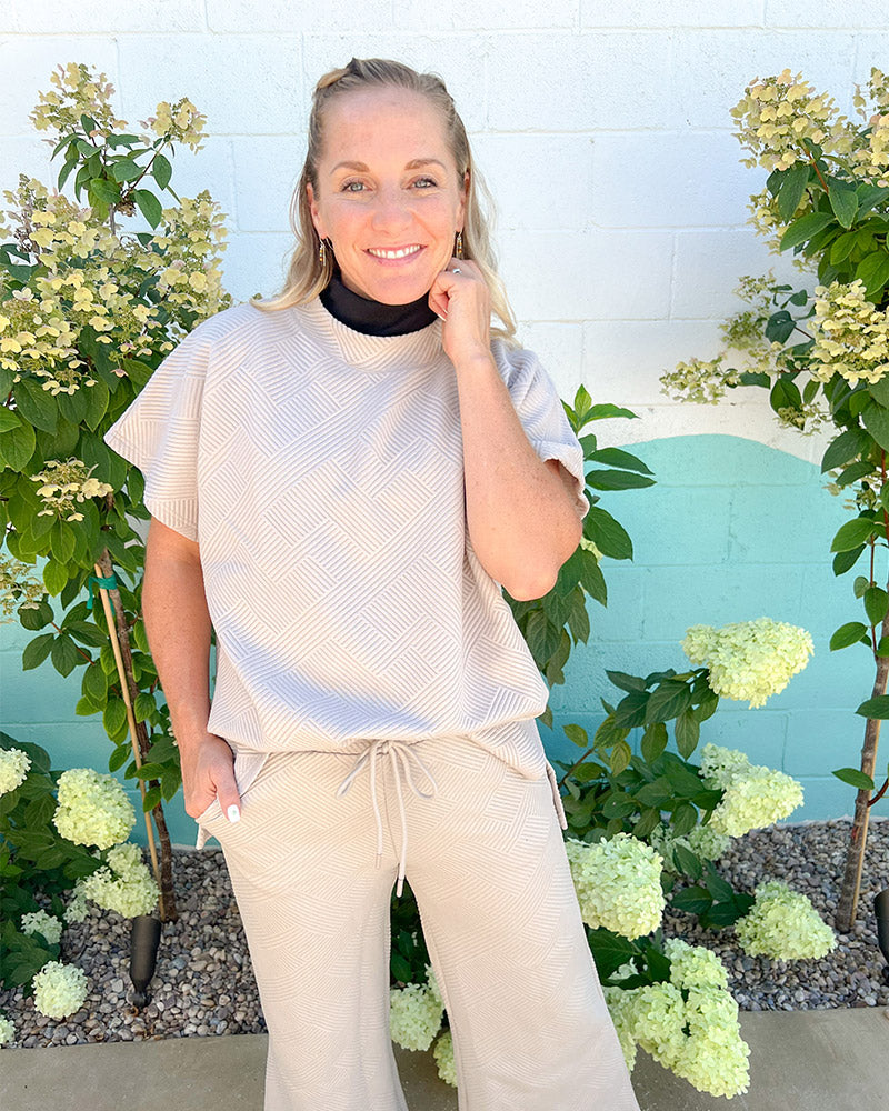 A woman wearing a beige top and wide leg pants, posing in a mock neck sleeveless front outfit.