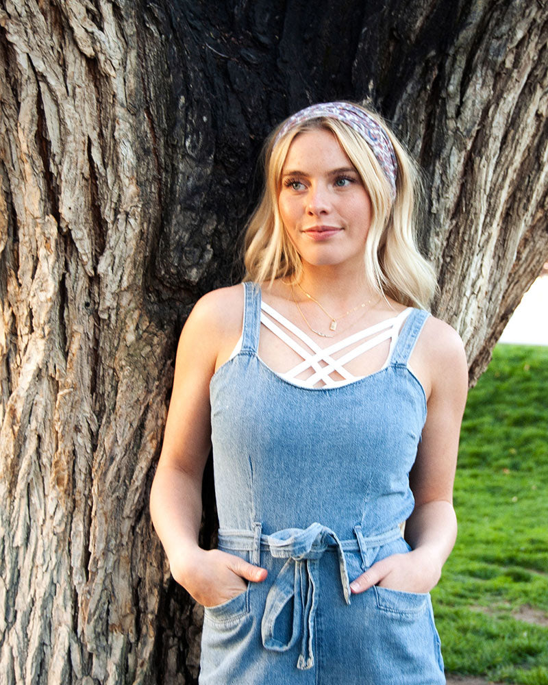 A blonde woman in a denim jumpsuit standing next to a tree, wearing a Strappy Criss Cross Cami Bralette.